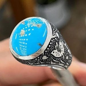 Könnte beinhalten: Ein silberner Ring mit einem großen, runden, helltürkisen Stein. Das Band des Rings weist komplizierte, dunkle, eingravierte Muster auf. Der Türkisstein hat kleine, goldfarbene Flecken. Der Ring wird von einem silbernen Werkzeug gehalten.