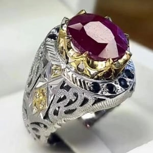 Op de afbeelding: Een zilveren ring met een ovale, dieprode edelsteen. De ring heeft ingewikkelde filigraan en gouden accenten rond de edelsteen en op de band. Kleine zwarte en gele stenen zijn in het ontwerp gezet. De ring wordt in een witte doos getoond.