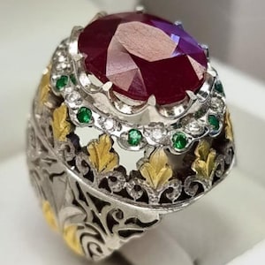 Puede incluir: Anillo de plata con un gran rubí ovalado como pieza central. El anillo presenta intrincados detalles de filigrana dorada y plateada, con pequeños acentos de esmeraldas y diamantes alrededor del rubí. Presentado en una caja de joyería blanca.