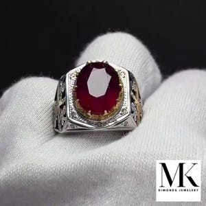 May include: A silver ring with a large, oval-cut ruby gemstone. The ruby is a deep red color and is set in a gold-colored bezel. The ring has intricate detailing on the band, with a decorative pattern. The ring is displayed on a white surface.