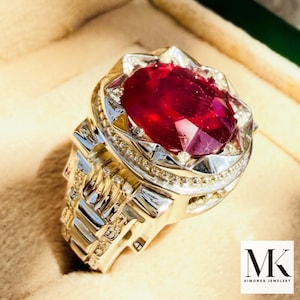 Könnte beinhalten: Ein auffälliger Ring mit einem großen, oval geschliffenen Rubin. Der Ring hat ein silberfarbenes Band mit aufwendigen Details und kleinen Akzentsteinen. Der Ring wird in einer beigen Schmuckschatulle präsentiert.