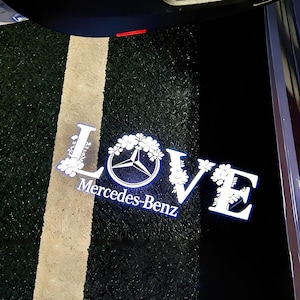 May include: A white neon sign with the word "LOVE" and the Mercedes-Benz logo, surrounded by floral accents. The sign is on a black and green textured surface with a beige stripe.