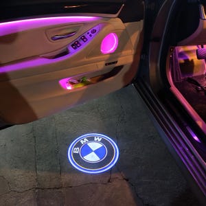 May include: A car door interior with purple ambient lighting. The BMW logo is projected onto the ground. The door panel is beige with control buttons and a speaker. The car door is open.