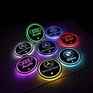 May include: A collection of illuminated car coasters featuring logos for Audi, Mercedes-Benz, and BMW. The coasters have a black base with the brand logo in the center, surrounded by a glowing ring of various colors like green, red, and blue.