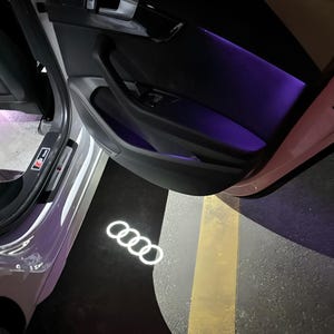 Puede incluir: Interior de la puerta de un coche con iluminación ambiental púrpura. El logotipo de Audi se proyecta en el suelo con luz blanca. La puerta está abierta, revelando el interior y el logotipo iluminado.
