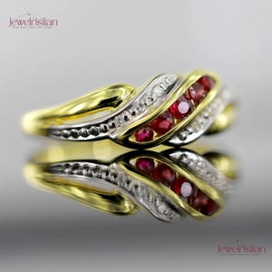May include: A gold and silver-toned ring with a wave design. The ring features a row of red gemstones and small white stones. The ring is reflected in the surface below. The text "Jewelrisitian" is visible in the image.