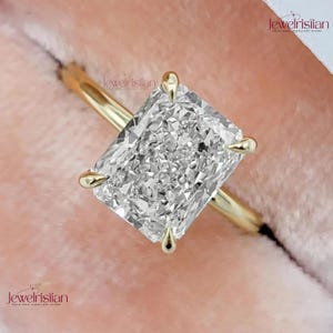 Puede incluir: Anillo de oro con un diamante de talla radiante. El diamante rectangular está sujeto por cuatro garras de oro. La banda del anillo también es de oro. El fondo rosa suave realza la elegancia del anillo.