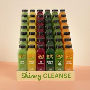 Puede incluir: Una exhibición de botellas de jugo en varios colores, incluyendo verde, amarillo, rojo y naranja, con tapas negras. Las botellas están etiquetadas con nombres como "Green Glow" y "Golden Defender". La exhibición está en una caja con el texto "Skinny Cleanse."