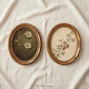 May include: Two oval-shaped framed floral prints. One features white flowers with green leaves against a dark background. The other displays light brown flowers and green leaves on a cream background. The frames are gold-colored.