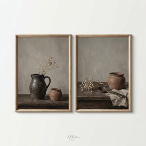 Könnte beinhalten: Zwei gerahmte Kunstdrucke mit Stillleben-Kompositionen. Ein Druck zeigt einen dunklen Krug und einen kleinen Topf mit getrockneten Blumen. Der andere Druck zeigt einen Terrakotta-Topf, einen Holzlöffel und ein Tuch mit getrockneten Blumen. Die Drucke haben eine neutrale Farbpalette.