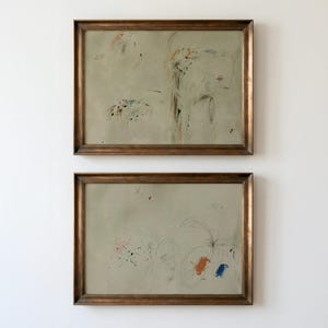 May include: Two framed abstract art pieces. Each piece features a light gray background with colorful brushstrokes and scribbled lines in shades of orange, blue, and black. The frames are a warm, dark brown.