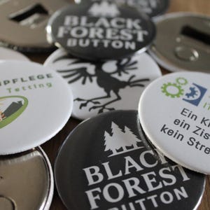Könnte beinhalten: Eine Sammlung runder Knöpfe und Flaschenöffner. Einige Knöpfe sind schwarz mit weißem Text "BLACK FOREST BUTTON". Andere Knöpfe haben Logos und Text, darunter "GARTENPFLEGE Traub & Tetting" und "Aichele IT Consulting".