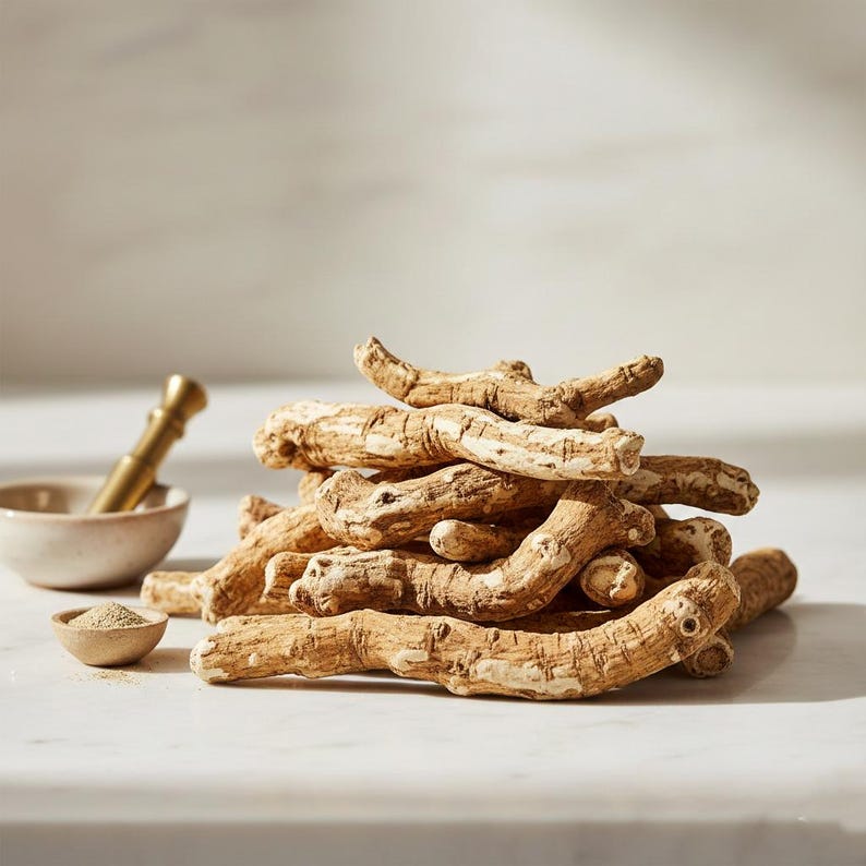 May include: A pile of dried, beige ginseng roots with a light-colored, textured appearance. A small, beige bowl with a golden pestle and a small wooden spoon with powder are in the background. The items are on a white surface.