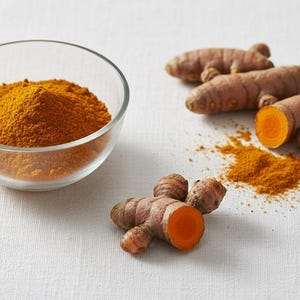 Peut inclure: Un bol en verre transparent rempli de poudre de curcuma orange vif est posé à côté de plusieurs racines de curcuma entières et tranchées. Les racines sont brunes avec un intérieur orange, et une petite pile de poudre est à proximité.