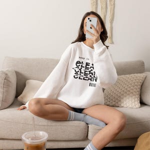 Peut inclure: Un sweat-shirt blanc à manches longues avec le texte noir "keep it" au-dessus d'un motif "CLEAN" répété et déformé, accompagné d'une petite icône de code-barres. Le vêtement présente un col rond, des poignets côtelés et une coupe décontractée.