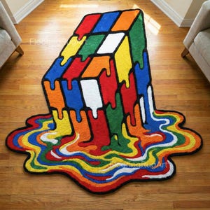 Op de afbeelding: Een handgemaakt vloerkleed in de vorm van een smeltende Rubik's Cube. Het kleed heeft blokken in rood, blauw, groen, geel, oranje en wit, met een zwarte omtrek. Het smelten creëert een uniek, artistiek ontwerp.