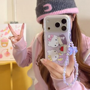 May include: Clear phone case with a purple backing featuring a cartoon design of two Hello Kitty characters. The case has a beaded wrist strap with a purple flower charm. The phone case is held up in front of a person.