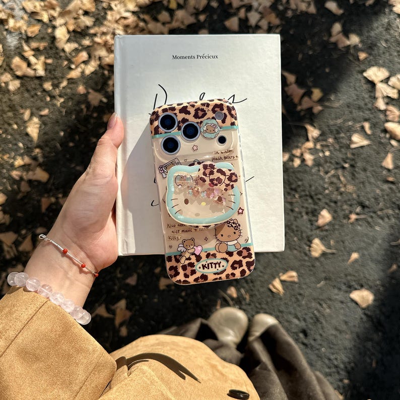 May include: A clear phone case with a leopard print design and a Hello Kitty theme. The case features a clear, liquid-filled Hello Kitty-shaped charm and the word "KITTY" at the bottom. The case is on top of a book.