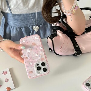 May include: A pink phone case with a clear border, featuring a pattern of small white cartoon characters and colorful stars. The case has a circular design in the center and a camera cutout. A pink purse and a silver heart necklace are also visible.