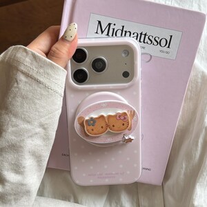 May include: Pale pink phone case with white spots and a Hello Kitty design. The case has a pop-up grip featuring two Hello Kitty characters. The text "seize the moment of happiness" is printed on the case.