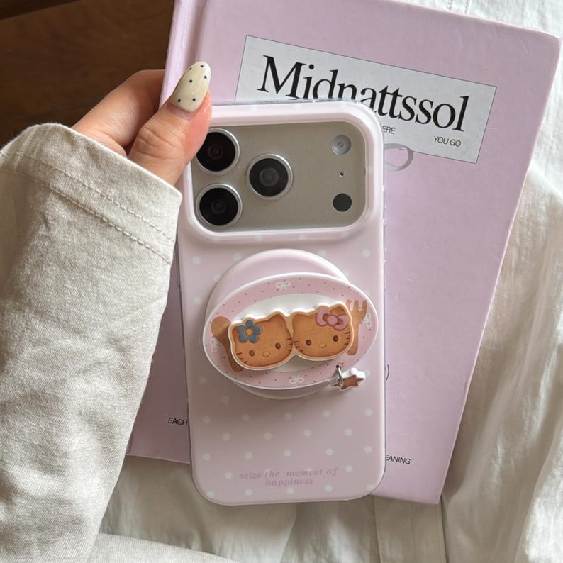 May include: Pale pink phone case with white spots and a Hello Kitty design. The case has a pop-up grip featuring two Hello Kitty characters. The text "seize the moment of happiness" is printed on the case.