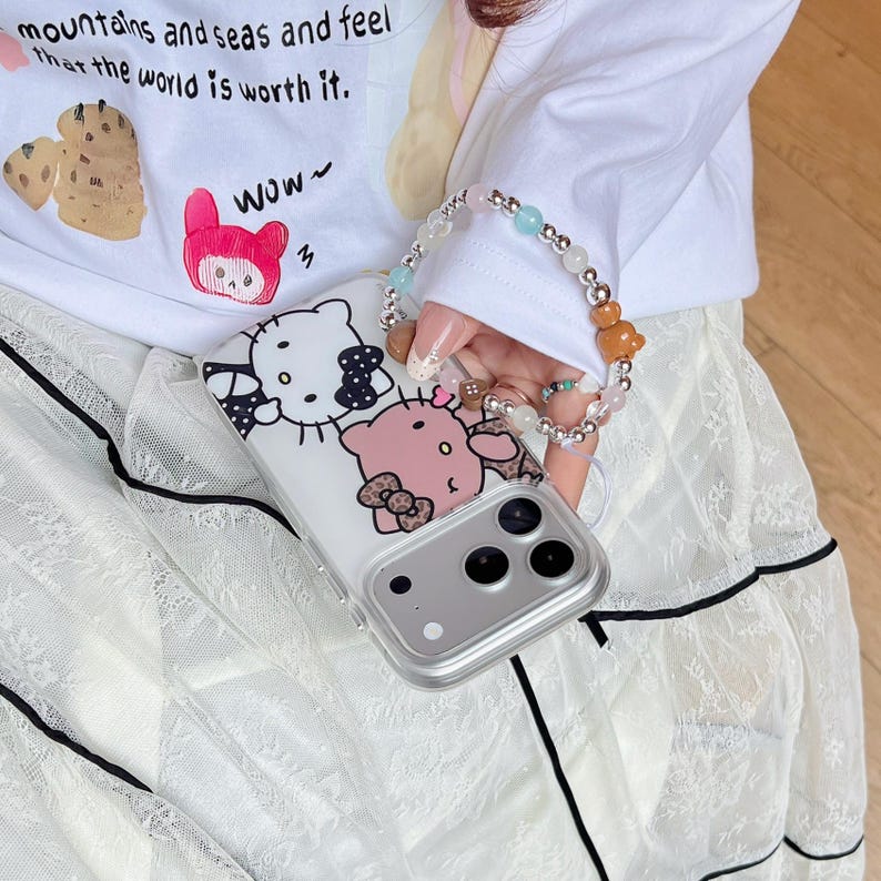 May include: A clear phone case featuring a cartoon design of two characters with bows. The case is held in front of a white long-sleeve shirt with text and a bracelet with colorful beads. The shirt has the text "mountains and seas and feel that the world is worth it."