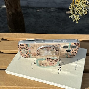 May include: Clear phone case with a leopard print design and a pop-up grip featuring a cartoon character. The case is on a book with the words "Moments Pr&eacute;cieux" and "Studio" visible.