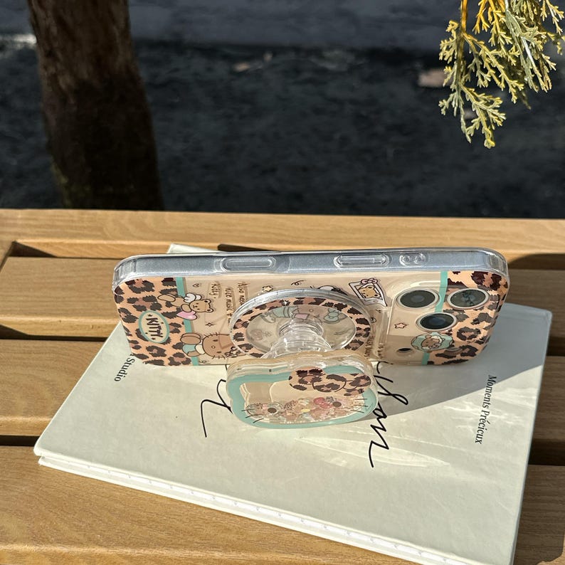 May include: Clear phone case with a leopard print design and a pop-up grip featuring a cartoon character. The case is on a book with the words "Moments Pr&eacute;cieux" and "Studio" visible.