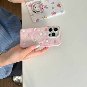 May include: Pink phone case with a clear ring holder and a matching pop-up phone grip. The case features a pattern of cartoon characters, stars, and dots. A small card with a Hello Kitty ring holder is also visible.