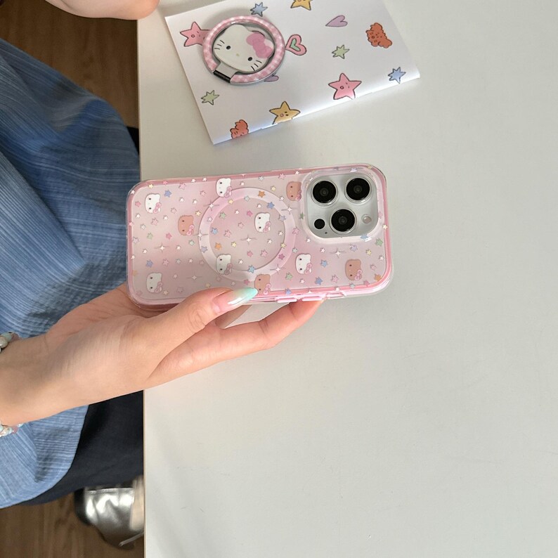 May include: Pink phone case with a clear ring holder and a matching pop-up phone grip. The case features a pattern of cartoon characters, stars, and dots. A small card with a Hello Kitty ring holder is also visible.