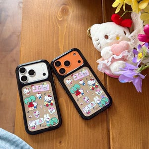 May include: Two black phone cases with a brown background featuring a repeating pattern of Hello Kitty characters, trees, and musical notes. One case has a white camera module, the other is orange. A Hello Kitty plushie and flowers are in the background.