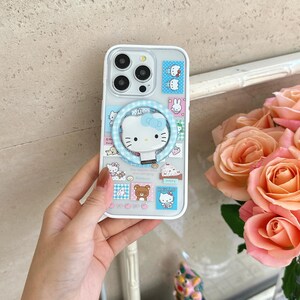 Puede incluir: Funda transparente para tel&eacute;fono con dise&ntilde;o de Hello Kitty, lazo azul y soporte de anillo. La funda tiene un borde blanco y est&aacute; decorada con varias im&aacute;genes tem&aacute;ticas de Hello Kitty en colores pastel.