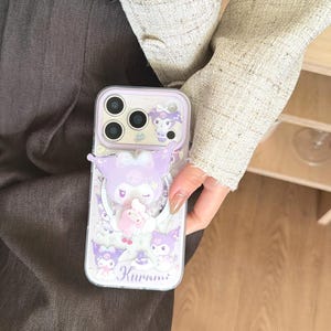 May include: A lavender phone case featuring a cartoon character with a bow and star accents. The case has a clear border and a pop-up design. The word "Kuromi" is printed on the bottom. The case is designed for a smartphone.