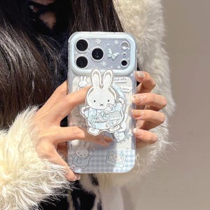 Może przedstawiać: Przezroczyste etui na telefon z jasnoniebieskim i białym wzorem Miffy. Etui posiada trójwymiarową białą postać Miffy, motyle i gwiazdki. Tło ma niebiesko-biały wzór w kratkę oraz tekst "Miffy" i "funny".