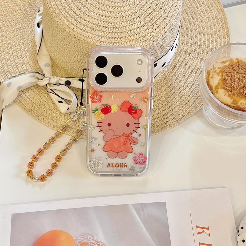 May include: Clear phone case with a Hello Kitty design. The case features a pink Hello Kitty character wearing a dress and a flower crown. The case has the word "Aloha" printed on it. A gold beaded wrist strap is attached to the case.