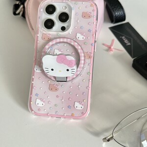 May include: Clear phone case with a pink and white Hello Kitty design. The case features a ring holder with a Hello Kitty face and a pink bow. The case is decorated with star and Hello Kitty face patterns.