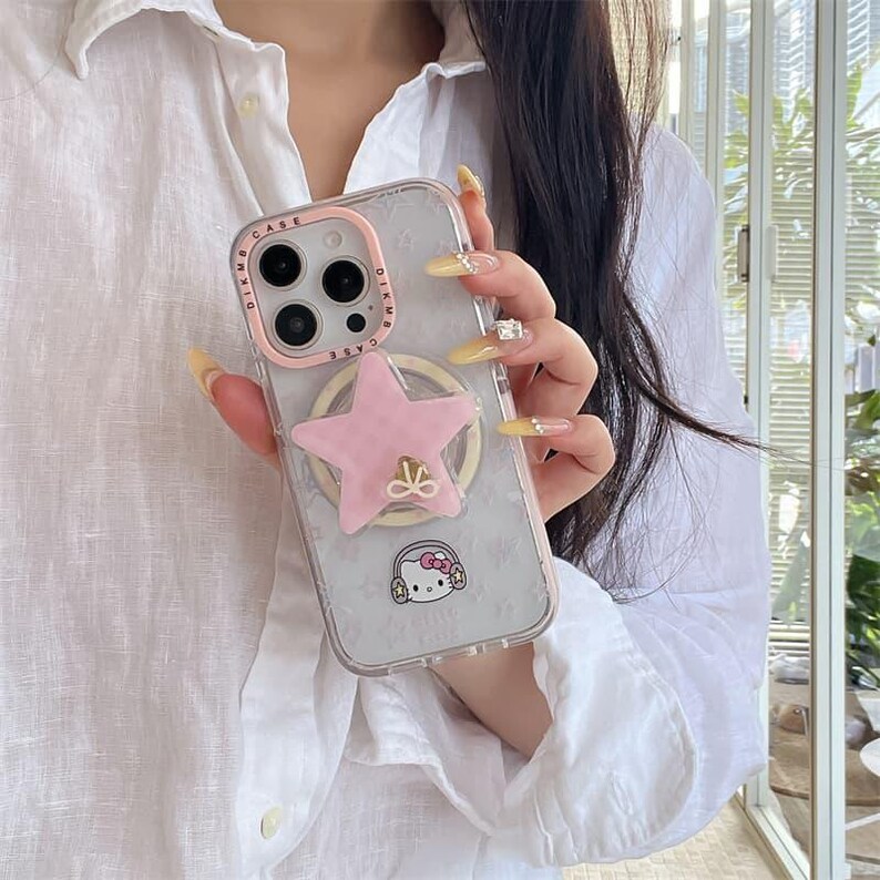 K&ouml;nnte beinhalten: Transparente Handyh&uuml;lle mit einem rosa sternf&ouml;rmigen Handyhalter und einem rosa Kamerarahmen. Die H&uuml;lle hat ein Hello Kitty-Motiv und ein dezentes Muster. Die H&uuml;lle ist f&uuml;r ein bestimmtes Telefonmodell konzipiert.