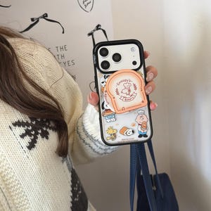 Może przedstawiać: Przezroczyste etui na telefon z czarną obwódką z Snoopy'm i przyjaciółmi. Etui posiada wysuwany uchwyt w kształcie kromki chleba z napisem "Snoopy's Bakery". Na etui wydrukowano różne postacie z kreskówek.
