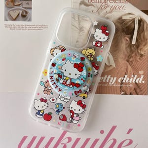 May include: Clear phone case featuring Hello Kitty and other cartoon characters. The case has a pop-up grip with a Hello Kitty design. The case is decorated with various images of Hello Kitty, flowers, and other cute elements.