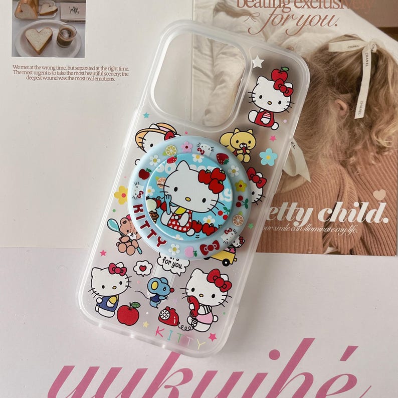 May include: Clear phone case featuring Hello Kitty and other cartoon characters. The case has a pop-up grip with a Hello Kitty design. The case is decorated with various images of Hello Kitty, flowers, and other cute elements.