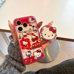 May include: Clear phone case with a red border and Hello Kitty graphics. The case features various Hello Kitty illustrations, including the character on a bus and holding strawberries. A matching pop socket with a Hello Kitty face is also visible.