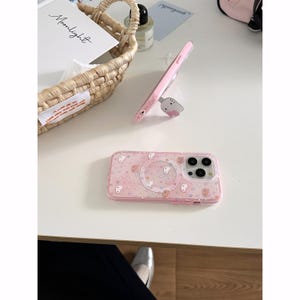 May include: Pink phone case with a bear pattern and a clear circular area. The case has a built-in stand with a cat design. A second pink phone case is also visible, along with a woven basket and a card that says "Moonlight."