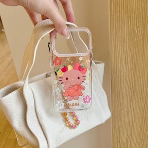 May include: Clear phone case featuring a pink cartoon character with a floral and fruit design. The case has a decorative charm and the word "Aloha" printed on it. The case is held up by a white purse strap.