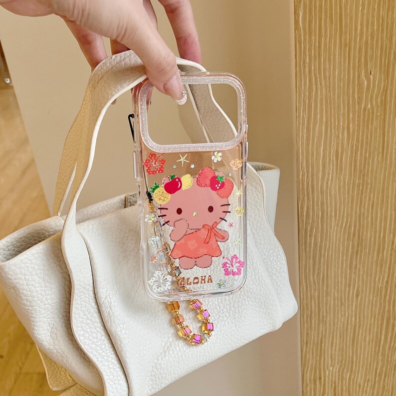 May include: Clear phone case featuring a pink cartoon character with a floral and fruit design. The case has a decorative charm and the word "Aloha" printed on it. The case is held up by a white purse strap.