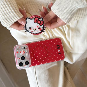 May include: Red phone case with white star pattern and a clear case with a Hello Kitty design. A matching Hello Kitty phone grip is held above the phone. The phone case has the text "I ❤️ KT".