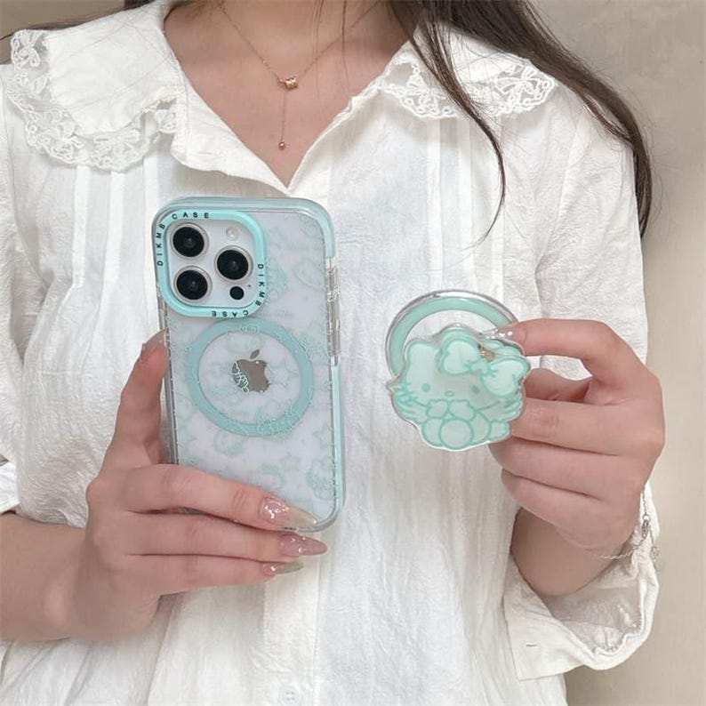 K&ouml;nnte beinhalten: Eine transparente Handyh&uuml;lle mit hellblauem Rand und einem passenden Pop-Socket. Die Handyh&uuml;lle hat ein hellblaues Apple-Logo und ein Muster aus kleinen Formen. Der Pop-Socket zeigt eine Zeichentrickfigur mit einer Schleife.