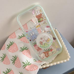 May include: Clear phone case with a mint green border, featuring cartoon characters and a glittery pop socket. The case rests on a strawberry-patterned cloth, with a light blue background.