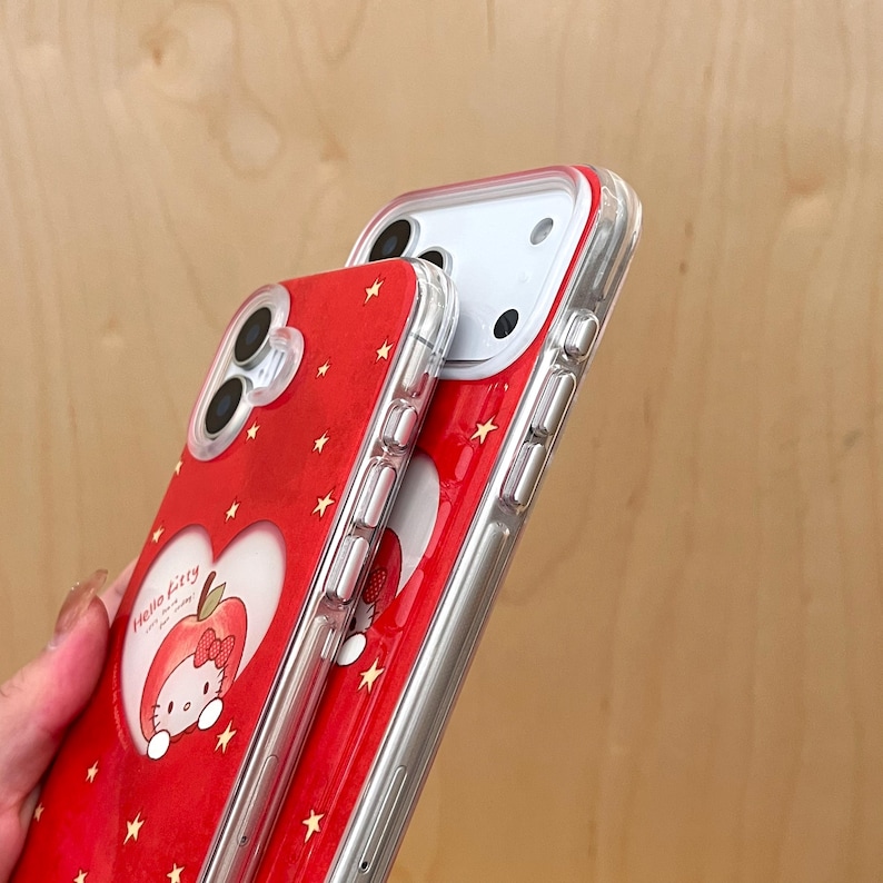 Puede incluir: Dos fundas rojas para tel&eacute;fono con borde transparente. Una funda presenta un dise&ntilde;o en forma de coraz&oacute;n con un gr&aacute;fico de manzana Hello Kitty y peque&ntilde;os detalles de estrellas. La otra funda es visible detr&aacute;s.
