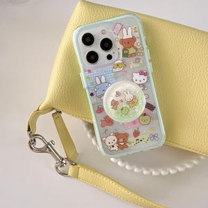 May include: A clear phone case with a light blue border, featuring cartoon characters and a pop-up grip. The case rests on a yellow purse with a matching strap and silver hardware. The phone case has a variety of cartoon characters.