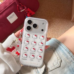 K&ouml;nnte beinhalten: Transparente Handyh&uuml;lle mit einem sich wiederholenden Muster von Hello Kitty-Gesichtern mit verschiedenen Ausdr&uuml;cken. Die H&uuml;lle hat ein silbernes Kameramodul und den Schriftzug "Hello Kitty". Eine rote Tasche und ein Herz-Charm sind ebenfalls zu sehen.